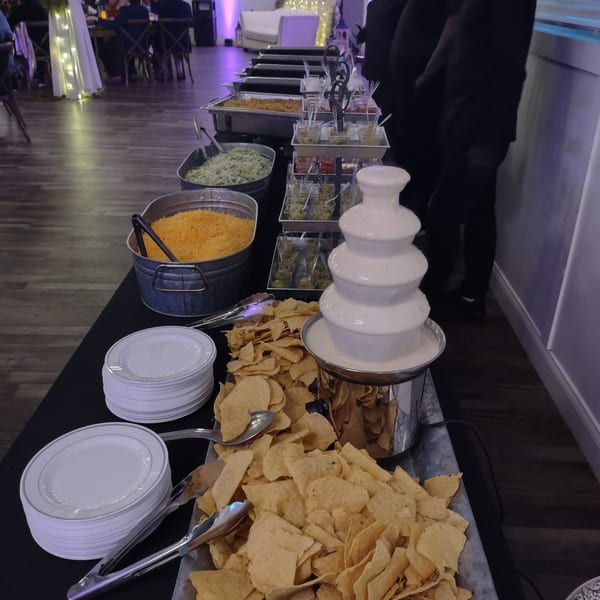A buffet table with a white cheese fountain, tortilla chips, shredded cheese, and other toppings.