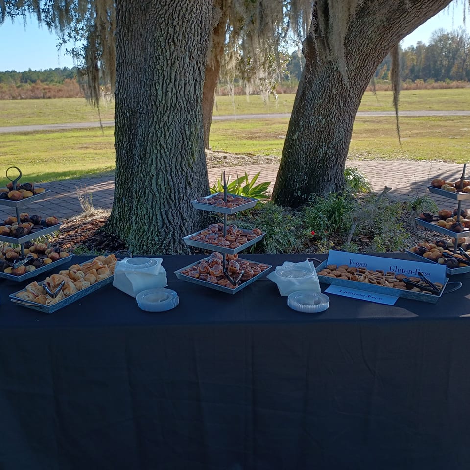 Outdoor buffet table with muffins, pastries, and cinnamon rolls under moss-draped trees.