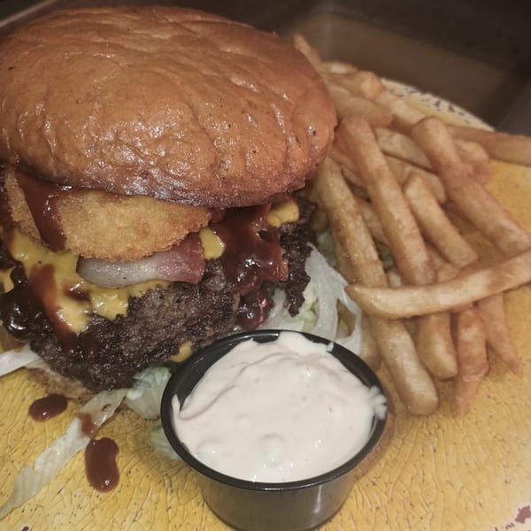 A Southern Taste Cafe burger with onion rings, bacon, cheese, and BBQ sauce, served with fries and ranch.