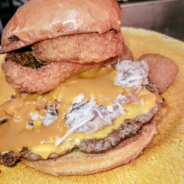 Southern Taste Cafe's burger with pulled pork, melted cheese, and onion rings on a yellow plate.